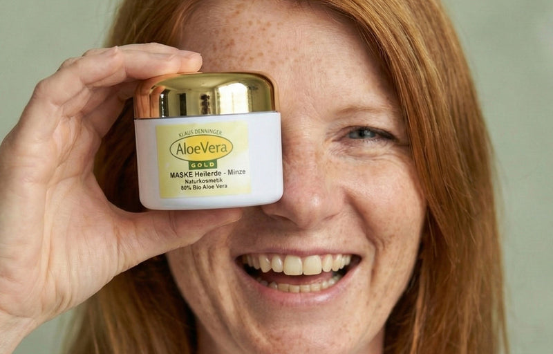 Eine herzlich lachende rothaarige Frau verdeckt ein Auge mit einem Kosmetiktiegel Aloe Vera Gold Maske Heilerde - Minze